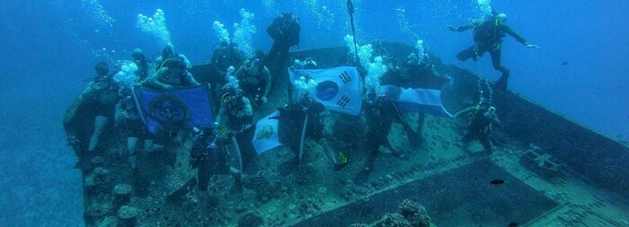 Divers hold up the Explosive Ordnance Disposal Mobile Unit 11 flag, along with the national flags of Peru, Republic of Korea, and The Netherlands, during a combined dive off the coast of Oahu, Hawaii during Exercise Rim of the Pacific (RIMPAC) 2024.