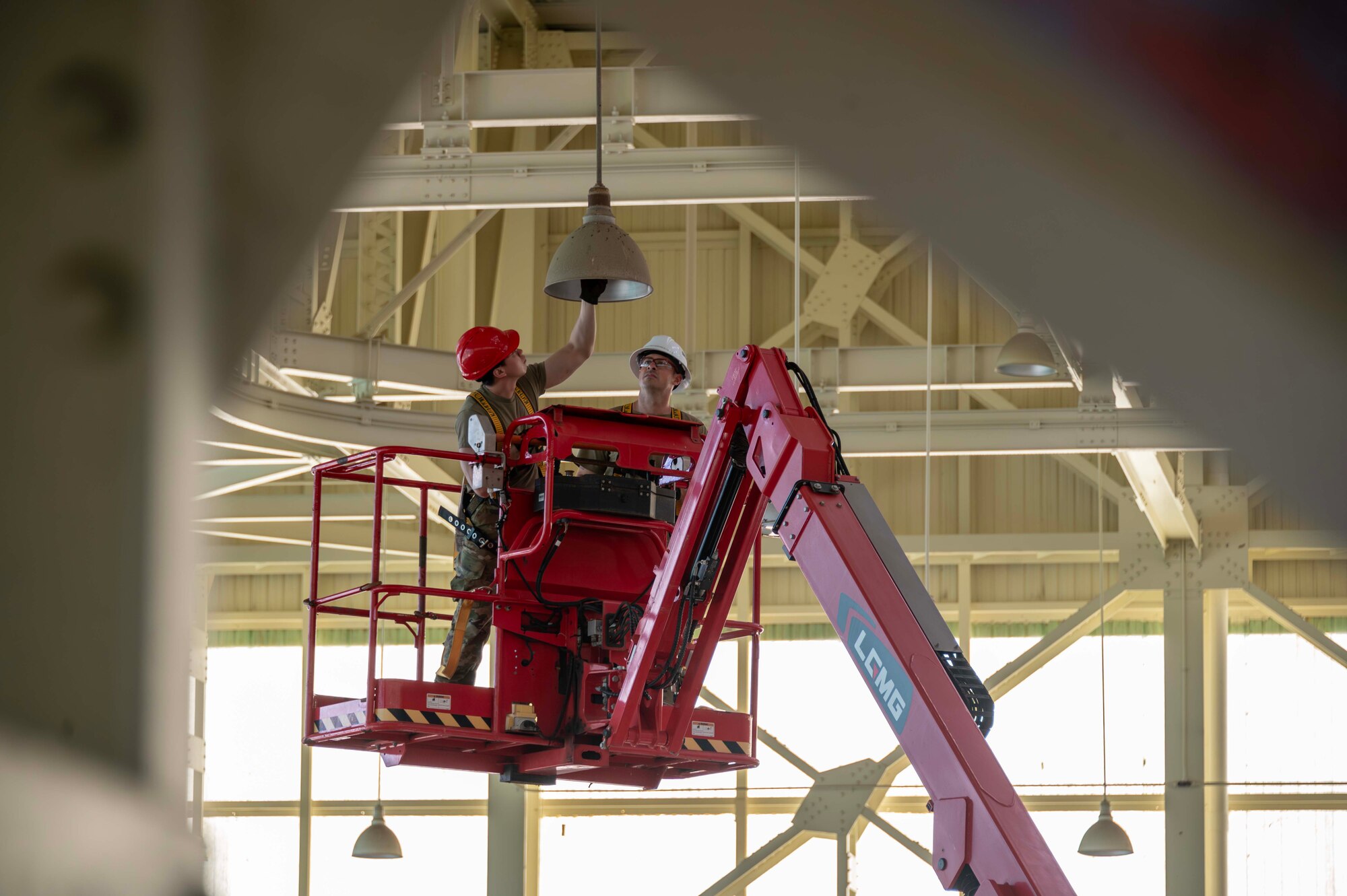 Two Airmen stand on a fixture to replace light bulbs