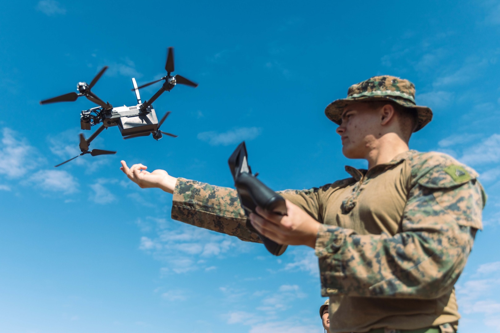 A Marine stands with an arm out while watching a drone flying close by.