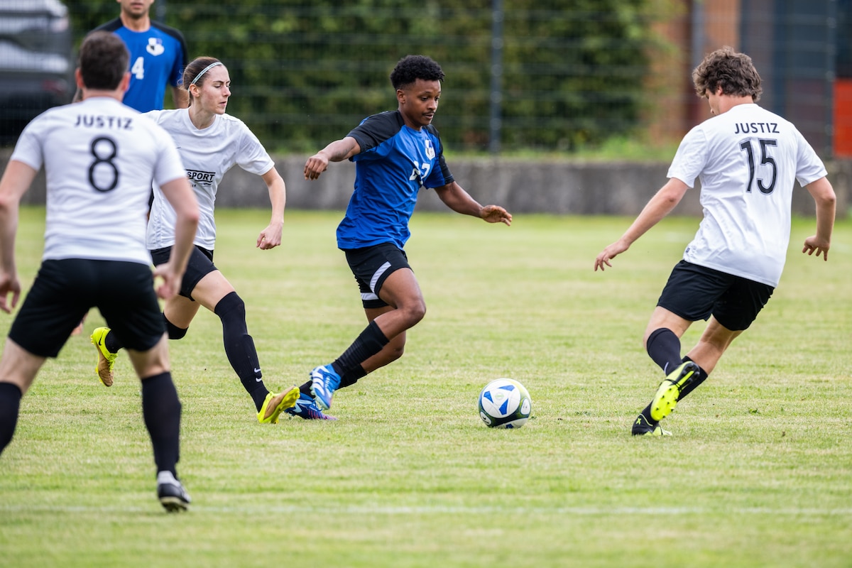 A soccer player in a blue jersey dribbles through two players in white jerseys.