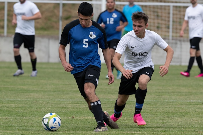 A soccer player in a blue jersey kicks a ball up a field as a player in a white jersey pursues him.