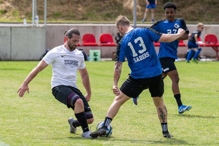 A soccer player in a blue kit attempts to kick a ball while a player in a white kit attempts to make a tackle