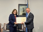 Two people (left: Ms. Joyce McKenna, right: Mr. Rob Landry) professionally dressed smiling and facing the camera both holding a paper award together in front of a couple chairs, a flag, and a picture on the wall.