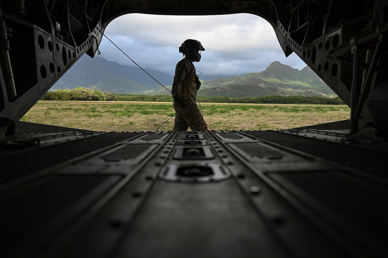 An airman wearing a helmet walks to the right outside the open deck of a military aircraft.