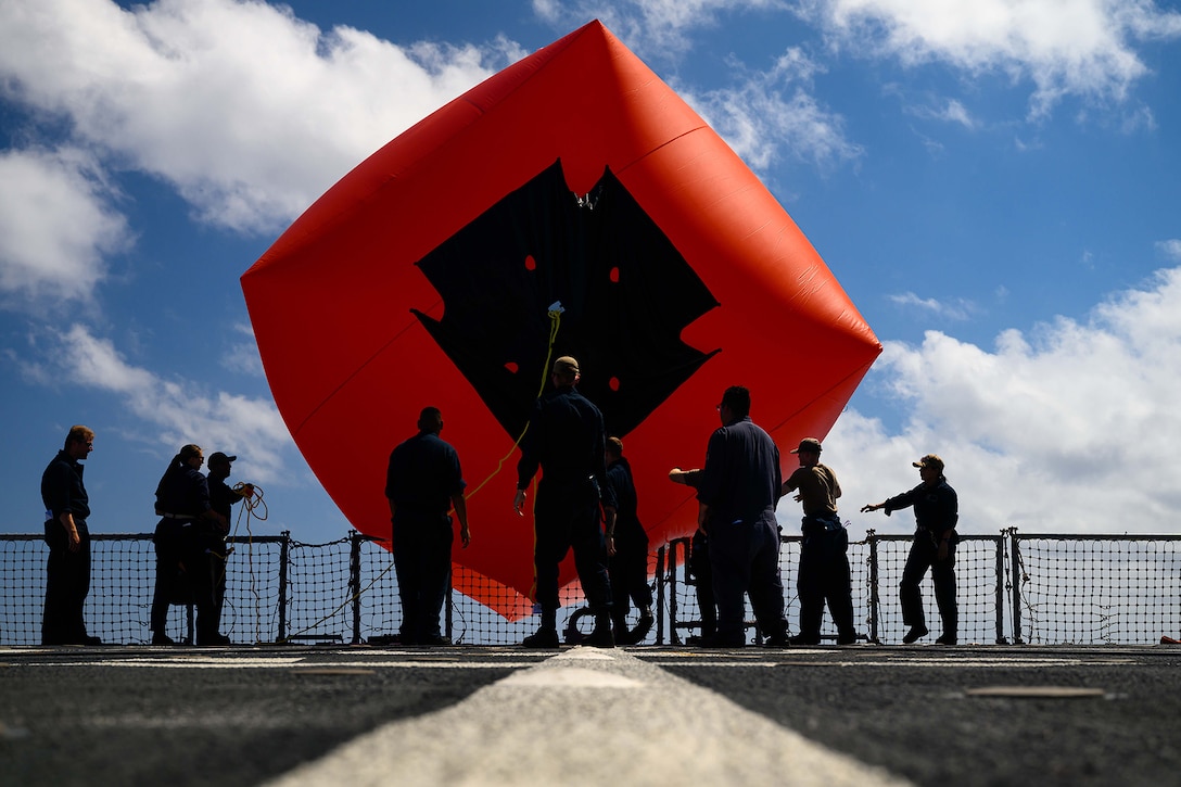 Sailors toss a large red inflatable target over the side of a Navy ship during daylight.