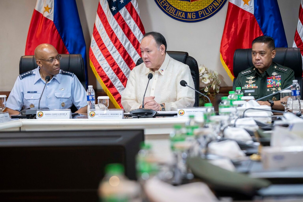 A U.S. service member sits at a table next to a person in civilian attire and another person wearing a foreign military uniform.