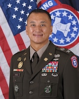 man wearing U.S. Army uniform standing in front of 2 flags.