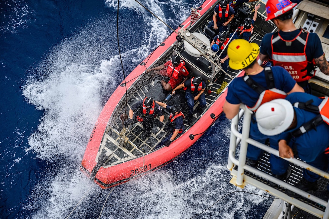 Three Coast Guardsmen aboard a ship look down as water splashes from fellow service members riding in a small rubber boat.