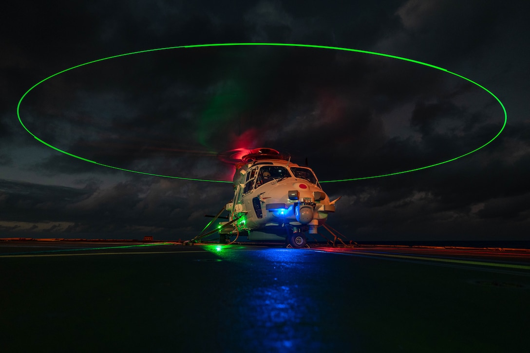 A helicopter sits on the flight deck of a ship in the dark illuminated by red, green and blue lights.