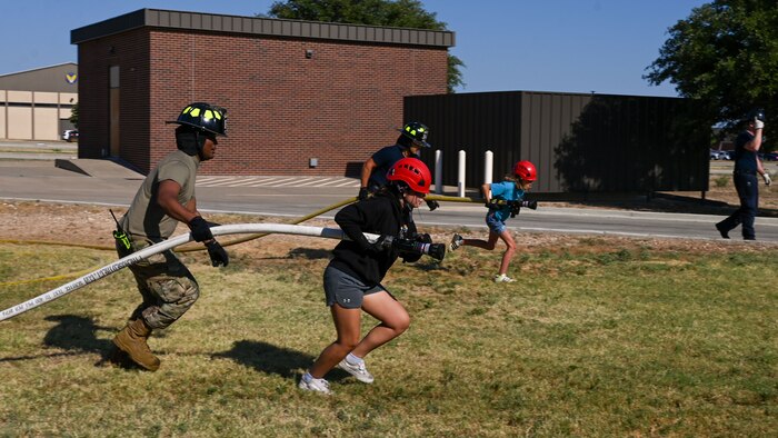 U.S. Air Force Airman First Class Wander Perez, Goodfellow Fire Department volunteer firefighter, assists Emmie in pulling the firehose to complete the Fire Hose Evolution during the fourth day of the 2024 Junior Firefighter Camp at the Goodfellow Fire Department, Goodfellow Air Force Base, Texas, July 11, 2024. Students practiced properly holding and dragging a fire hose, using its pressure to knock over a basketball and cone, and participated in a building search and rescue activity. (U.S. Air Force photo by Airman 1st Class Evelyn J. D’Errico)