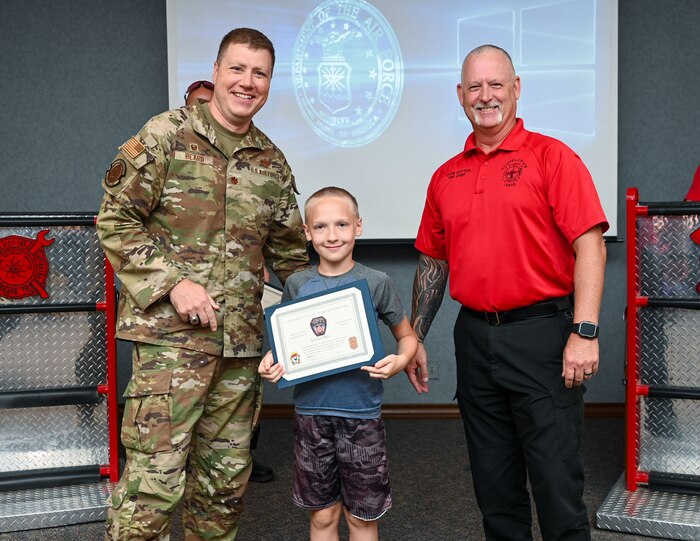 U.S. Air Force Maj. Levi Beard, 17th Civil Engineer Squadron commander, and Kevin Cotton, Goodfellow Fire Department fire chief, present Elijah, 11, with his graduation certificate during the 2024 Junior Firefighter camp at the Louis F. Garland Department of Defense Fire Academy, Goodfellow Air Force Base, Texas, July 12, 2024. After a week of different interactive lessons, the students earned the title of junior firefighters. (U.S. Air Force photo by Airman 1st Class Evelyn J. D’Errico)