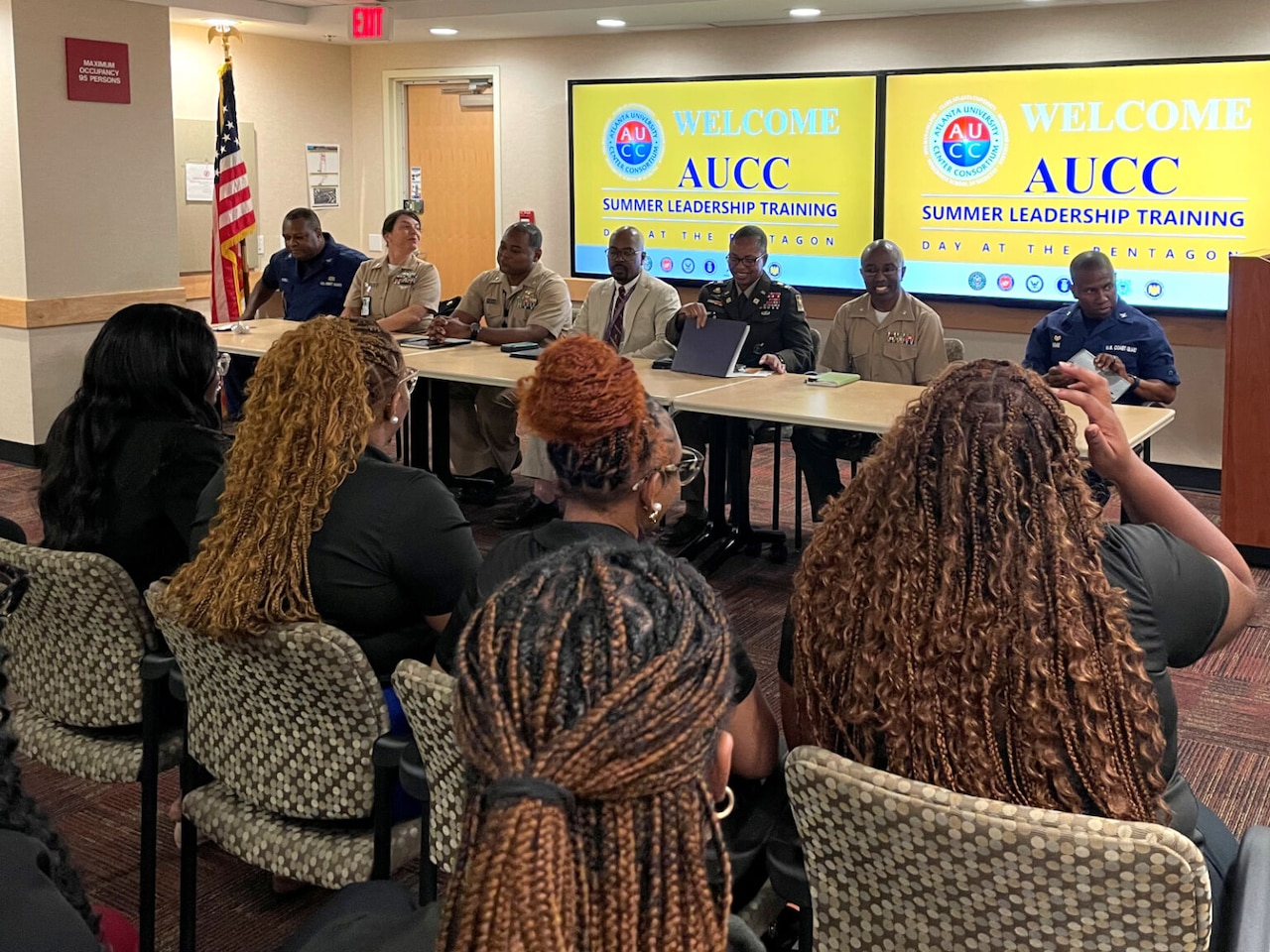 Seven individuals sit at a table. Behind them, screens say, "AUCC Summer Leadership Program." Young people are seated in front of them.