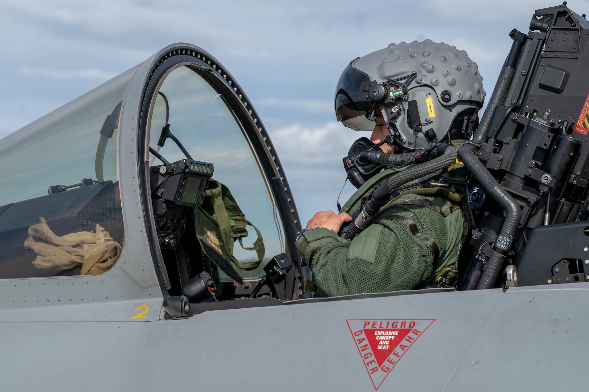 A service members from the Spanish Air Force sits in the cockpit of a Eurofighter Typhoon aircraft with his helmet on and the canopy of the aircraft open.