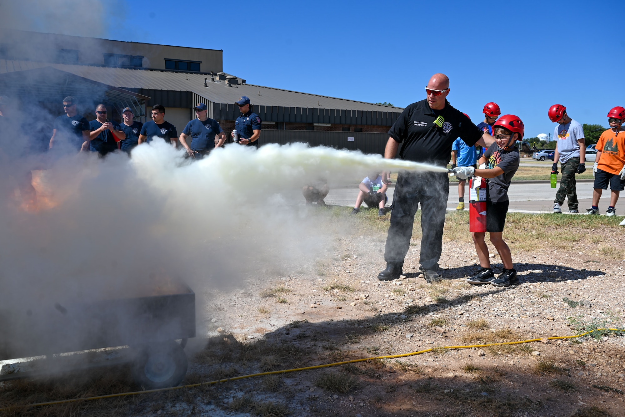 Brad Keibler, Goodfellow Fire Department fire inspector, helps Angel, 10, put out a propane fire with a fire extinguisher during the third day of the 2024 Junior Firefighter Camp at the Goodfellow Fire Department, Goodfellow Air Force Base, Texas, July 10, 2024. Keibler taught the junior firefighters the Pass, Aim, Squeeze and Sweep (PASS)  method of using a fire extinguisher. (U.S. Air Force photo by Airman 1st Class Evelyn J. D’Errico)