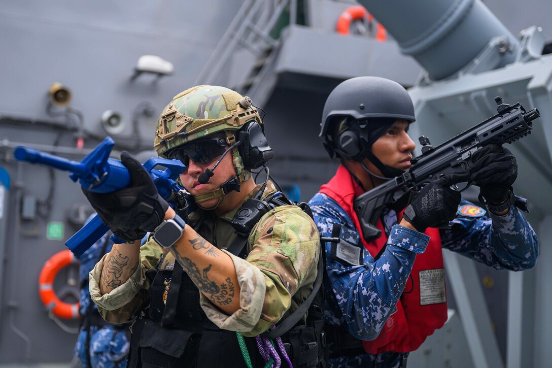Two soldiers on the deck of a ship and holding automatic weapons aim to either side.