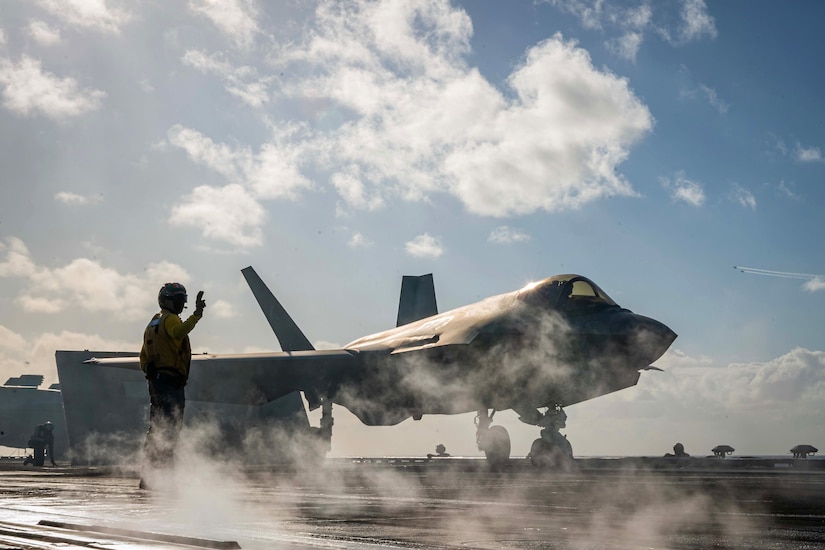 A sailor signals the pilot of a jet on a deck of a ship with smoke in front and clouds above.