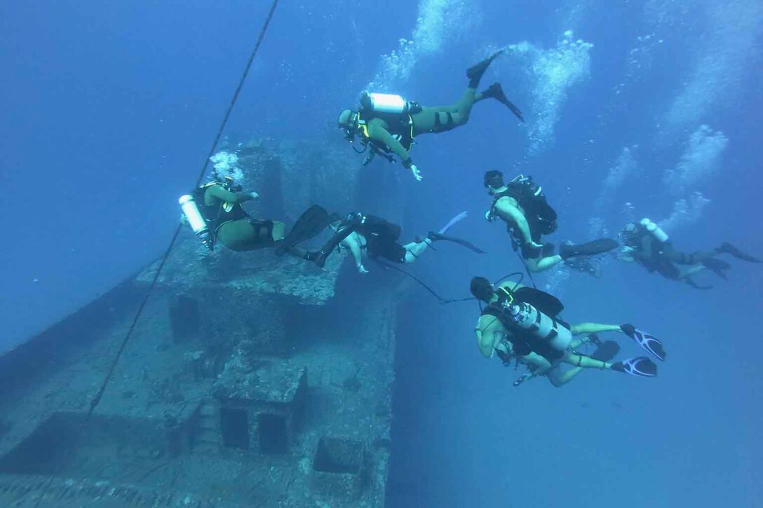 Service members wearing scuba gear dive together above the remains of a wrecked ship.