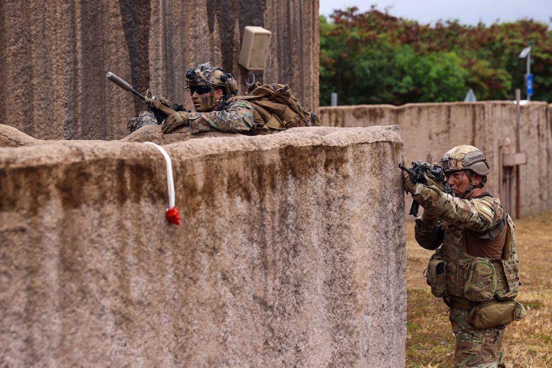 One Marine stands behind a wall looking ahead while another Marine stands next to the wall pointing a weapon.