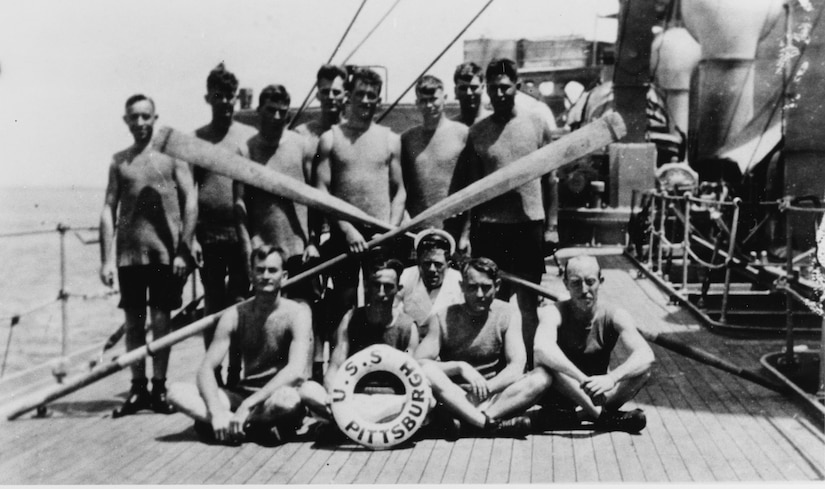 Two rows of men pose for a photo with large oars on the deck of a ship.