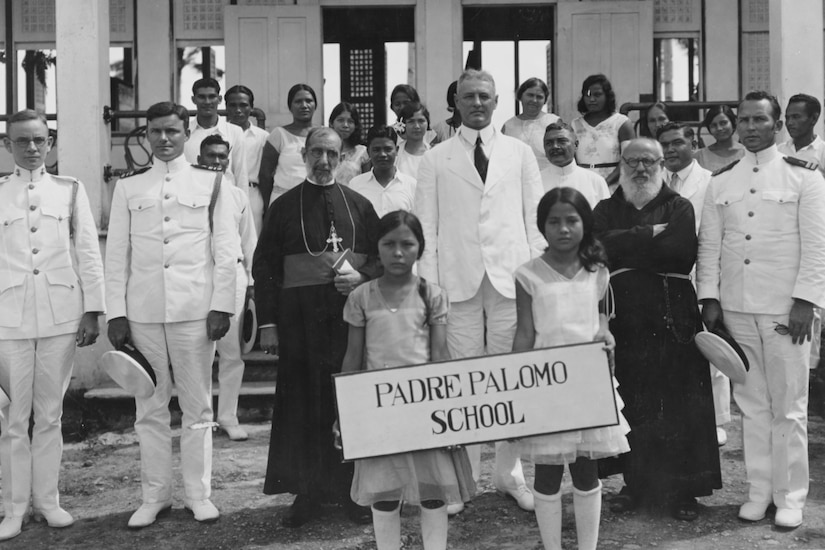 A group stands in front of a building posing for a photo. Two children hold a sign that says, “Padre Palomo School.”