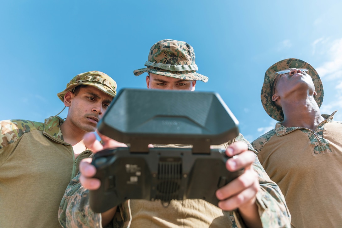 A service member looks up at the sky as two others look at a tablet as seen from below.