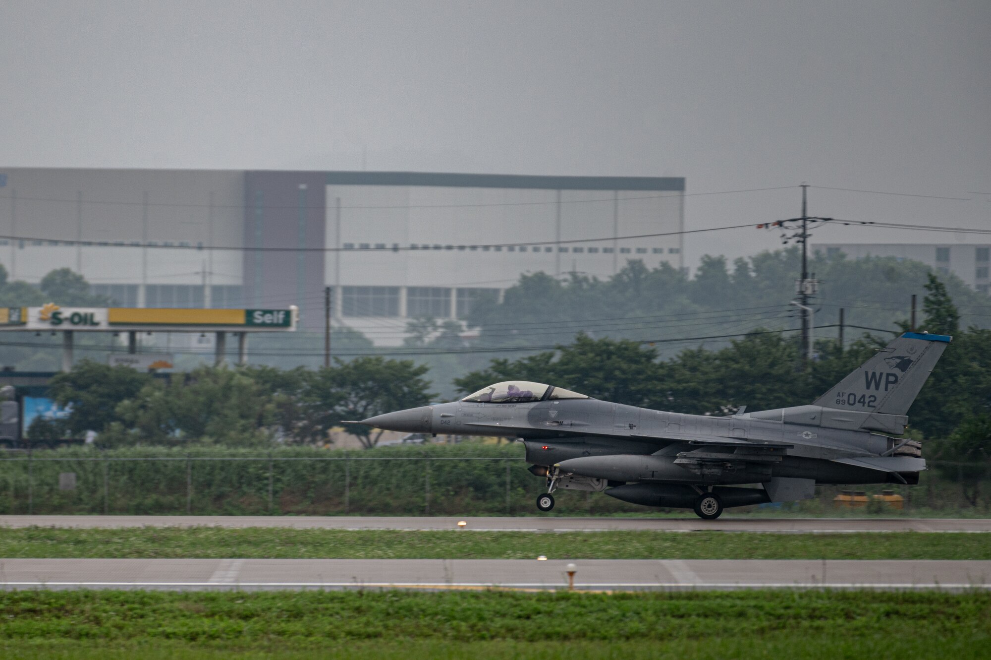 A gray F-16 fighter jet with Kunsan AB WP designation on its tail lands on a runway, viewed from the side profile with the nose pointing left, with green grass and a gray sky. There is a large gray building and trees in the background.