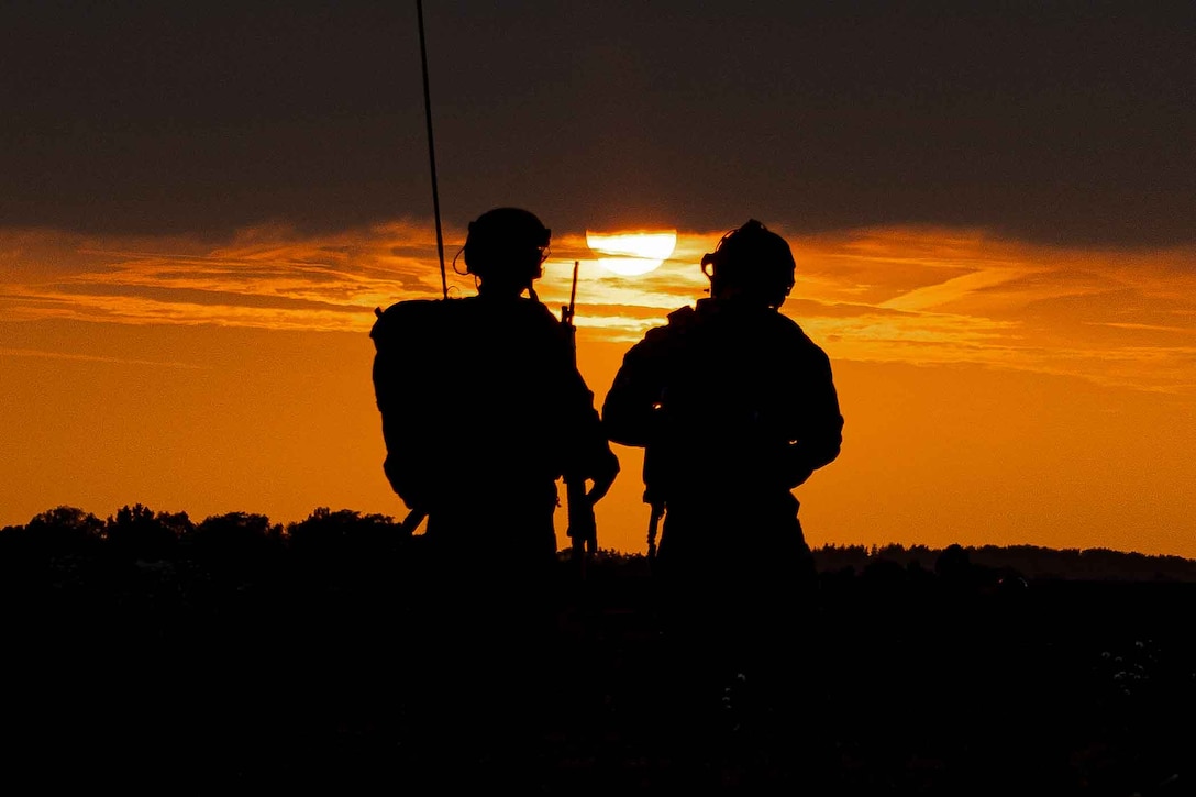 Two service members are silhouetted against an orange sky as they stand and look at the sun.