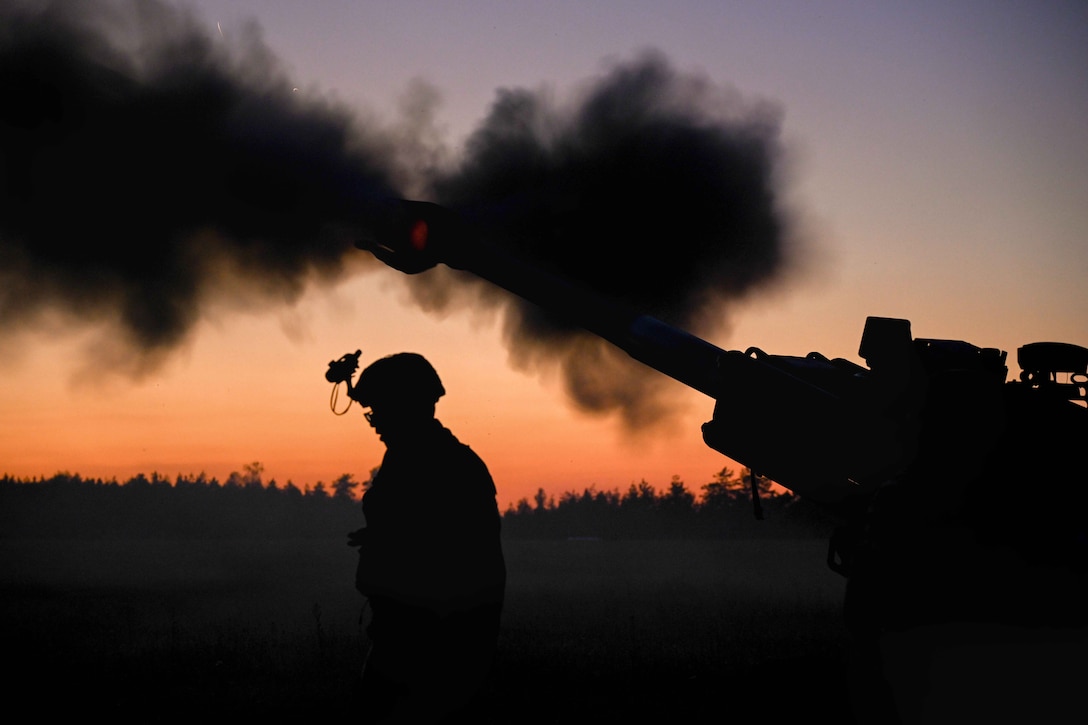 A soldier is silhouetted against an orange sky while standing close to a howitzer as it fires a rocket at twilight in a wooded area.