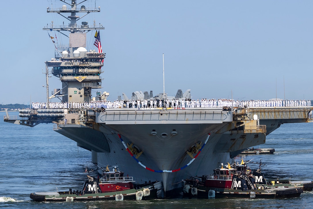 Sailors stand on the flight deck of a Navy ship as it returns to port during daylight.