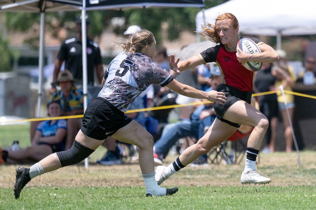 An athlete reaches for another athlete running with a rugby ball as spectators watch from the sideline.