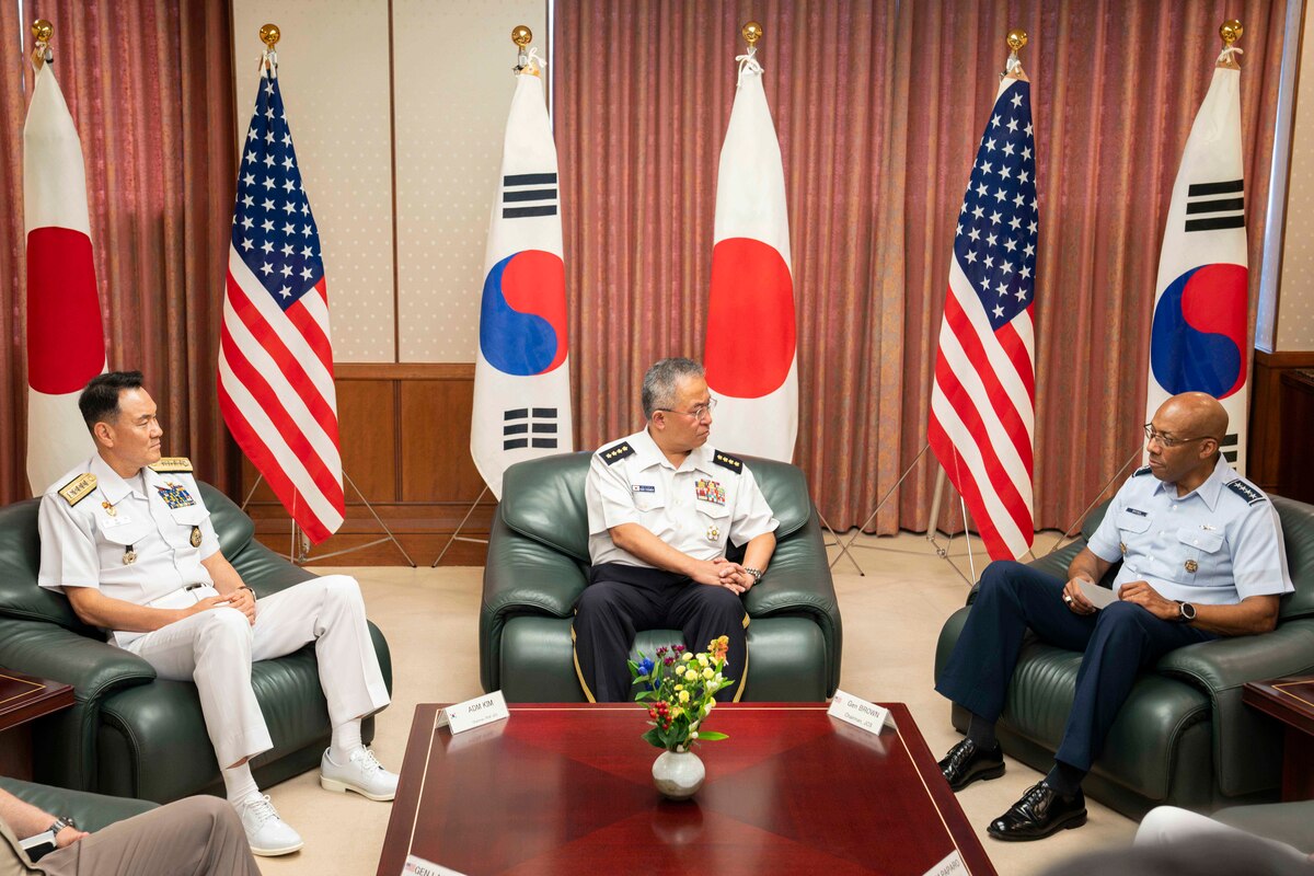 A U.S. service member is seated next to two foreign service members with U.S., Japanese and South Korean flags in the background.