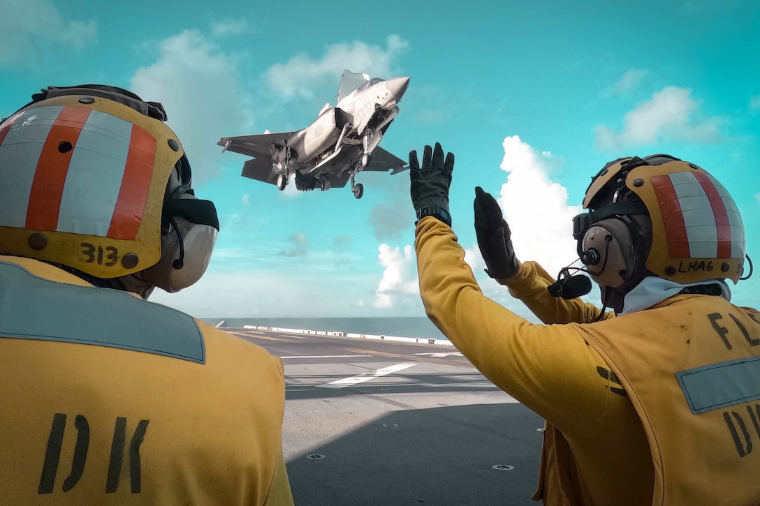 A sailor signals to an airborne aircraft as it prepares to land on the flight deck of a ship as a fellow sailor watches.