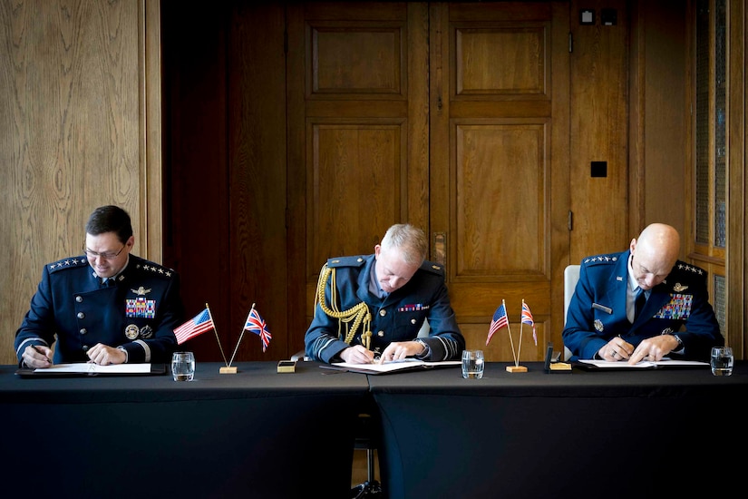 Three military officers sit at a long table and sign documents.