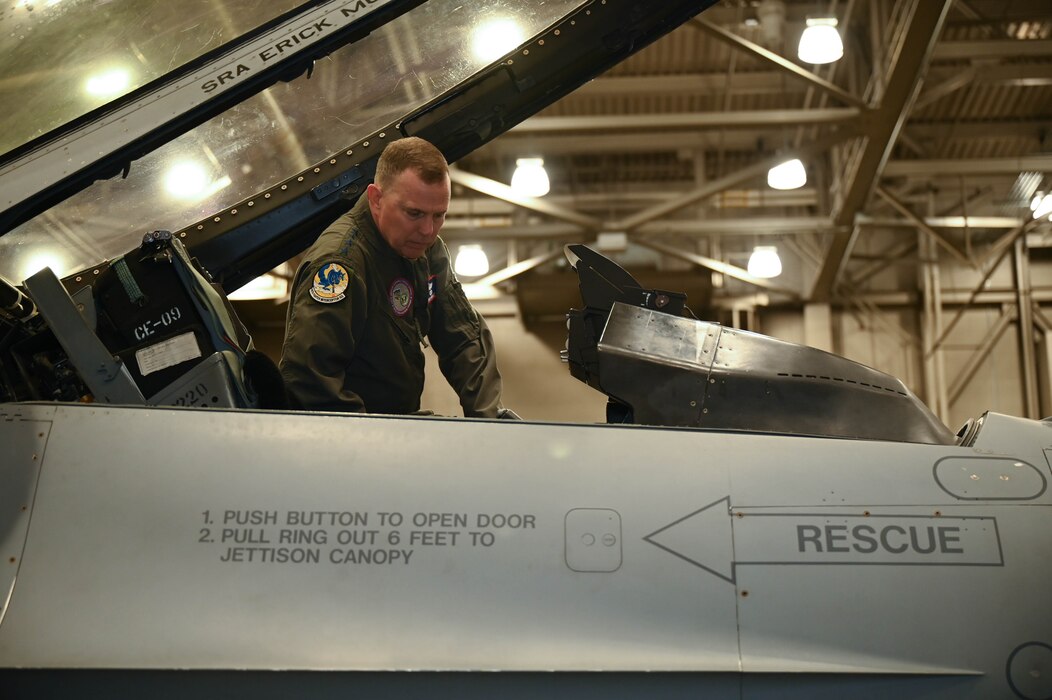 Gen. Guillot looks at the cockpit of an F-16 aircraft inside a lit hangar.