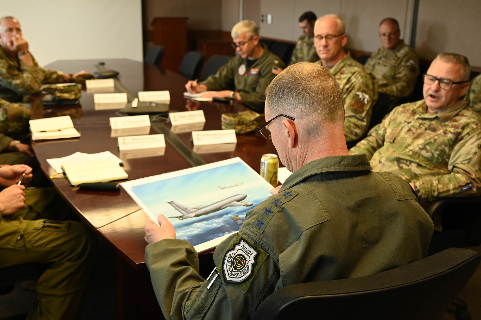 Gen. Guillot sits at the head of a table looking at a picture of a KC-135 tanker surrounded by 9 other people.
