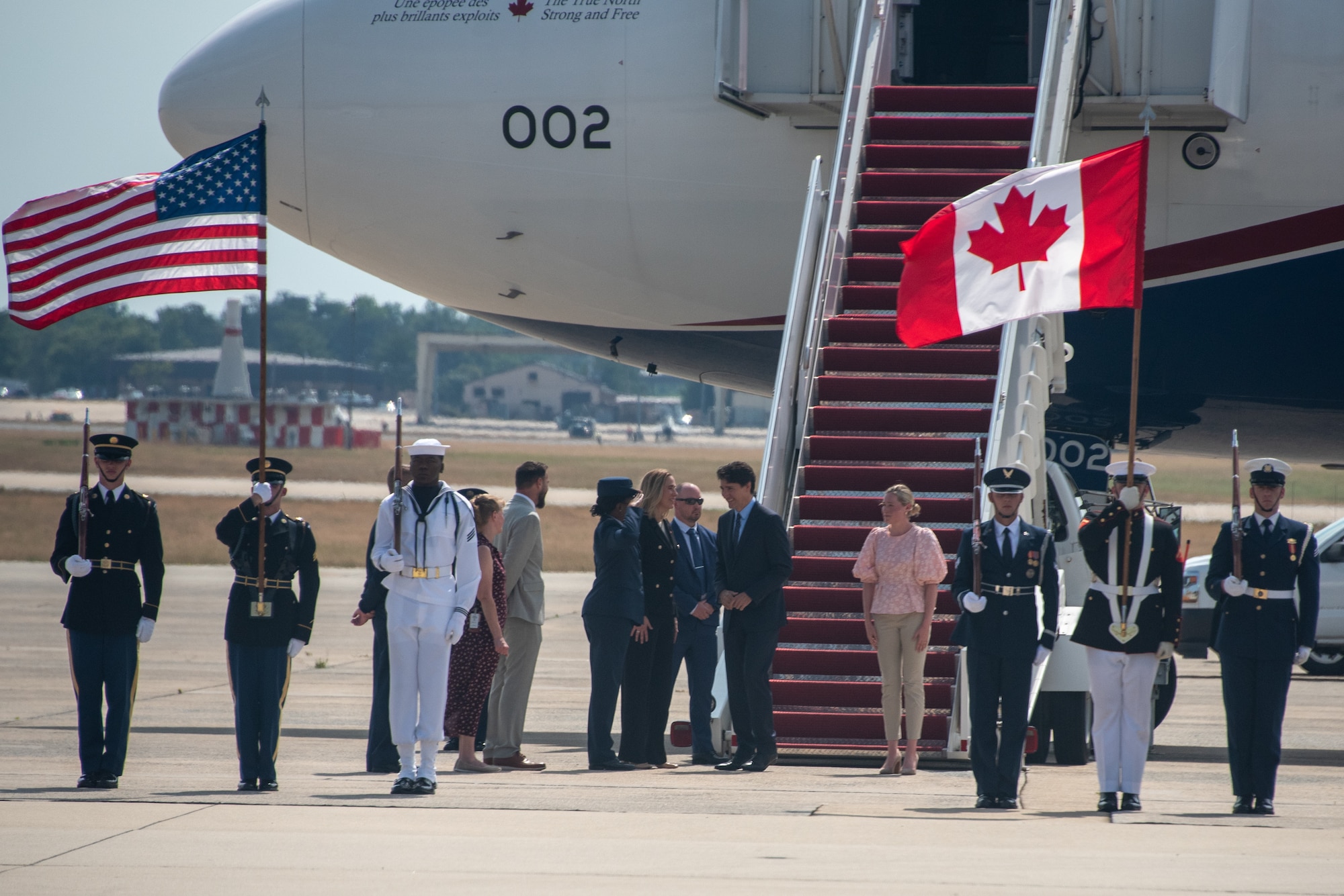 Men and women stand in front of a stair truck going up to a plane and next to service members holding up the United States and Canada flag.