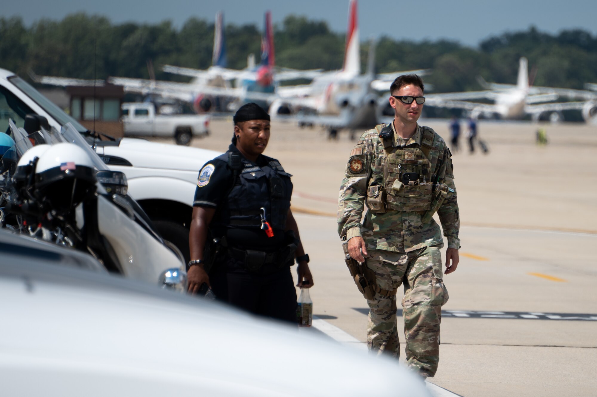 Two people walk side by side outside on a flightline in front of white vehicles.