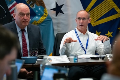 Two men sit at a table and the one on the right is speaking.