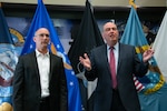 Two men stand in front of United States armed forces flags.