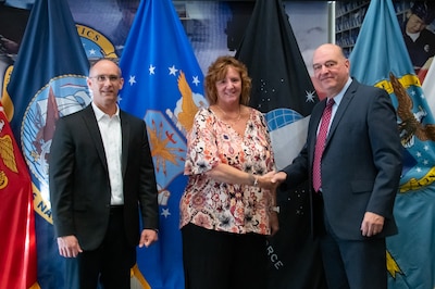 Two men and a woman stand in front of United States armed forces flags shaking hands.