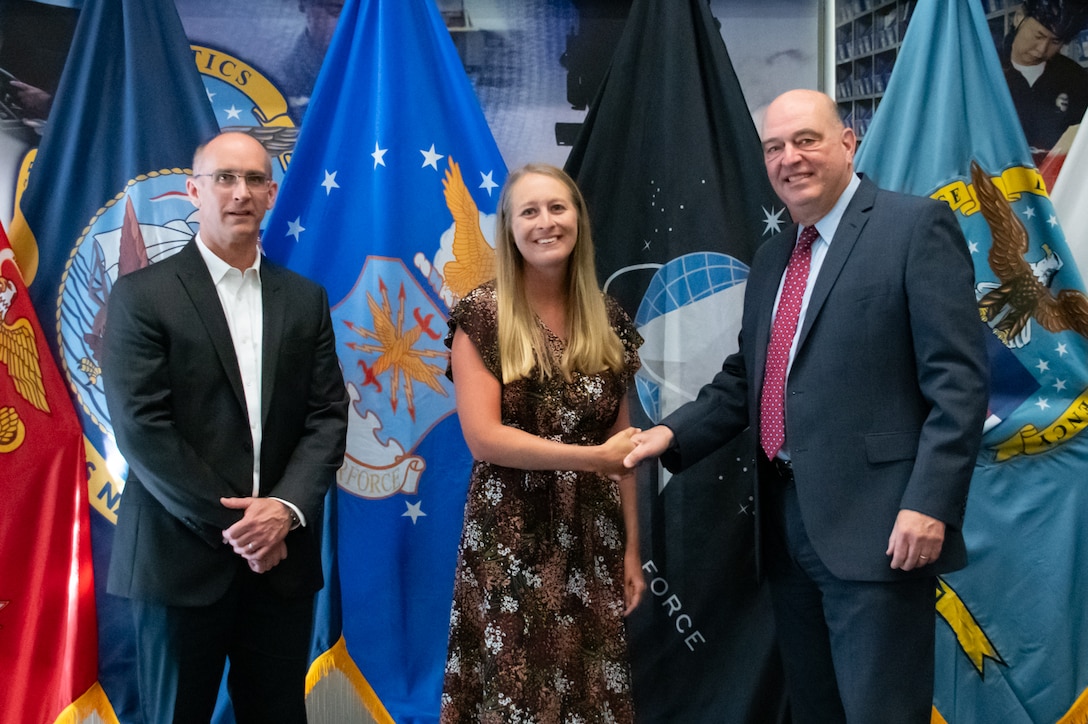 Two men and a woman stand in front of United States armed forces flags shaking hands.