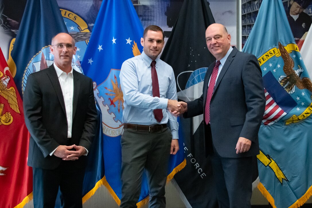 Three men stand in front of United States armed forces flags shaking hands.
