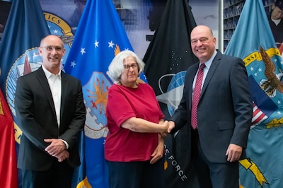 Two men and a woman stand in front of United States armed forces flags shaking hands.