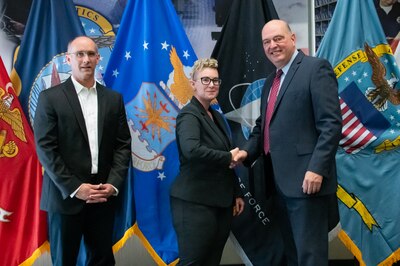 Two men and a woman stand in front of United States armed forces flags shaking hands.