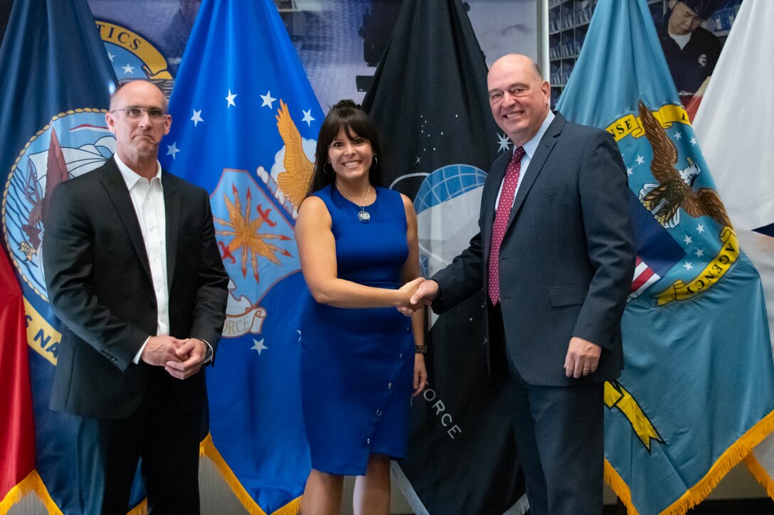 Three men stand in front of United States armed forces flags shaking hands.