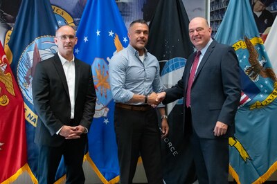 Three men stand in front of United States armed forces flags shaking hands.