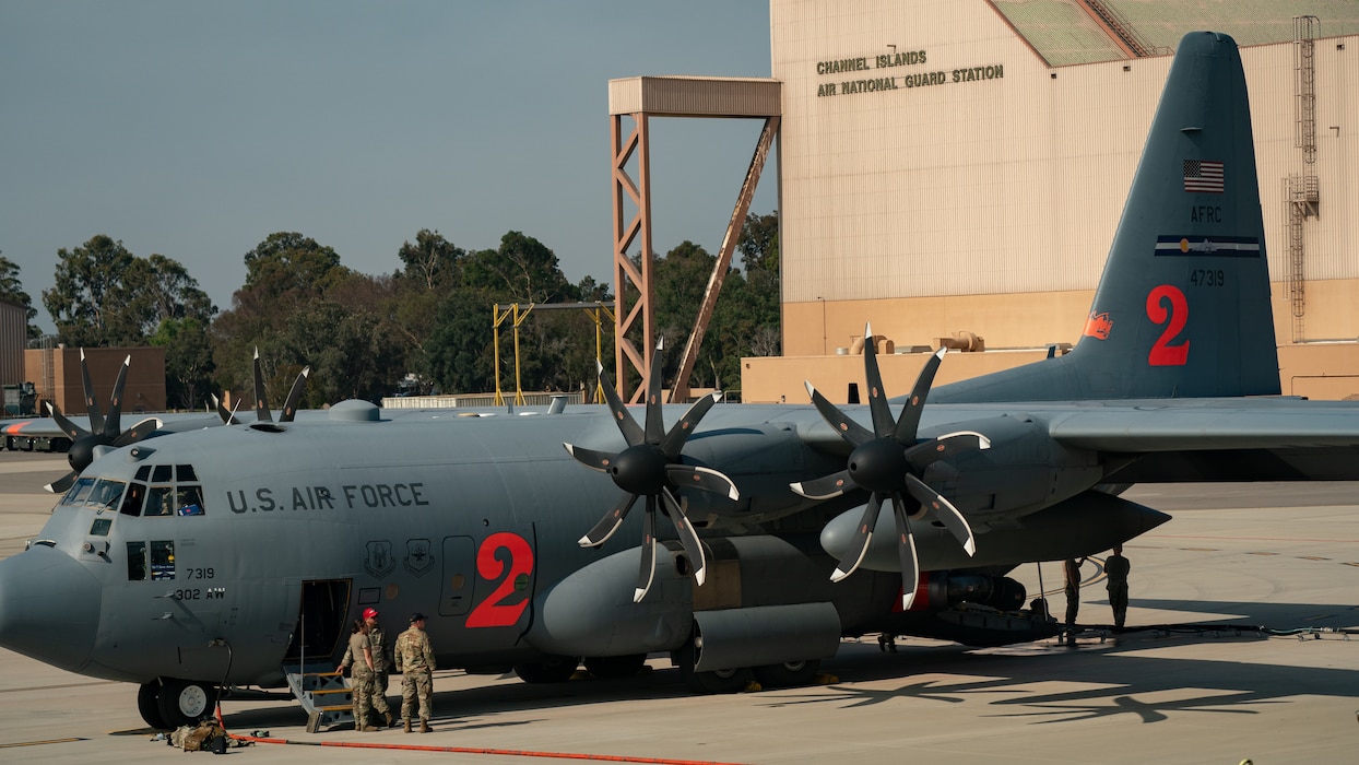 A side profile of parked MAFFS Air Force Reserve C-130 faces away from a giant military hangar as two Air Force Reserve Airmen exit the aircraft.