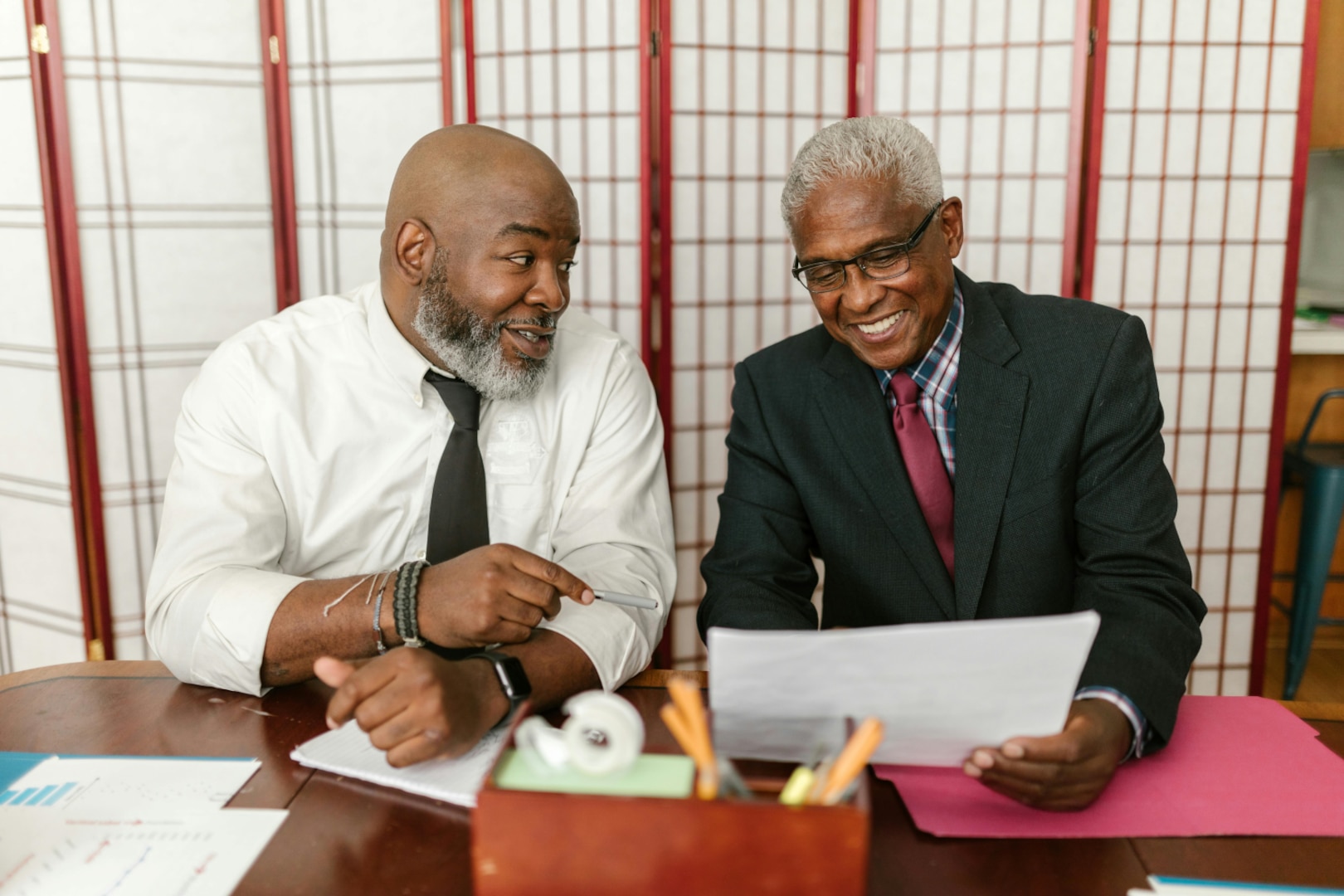 Two men wearing suits sit at a desk and look at a document.