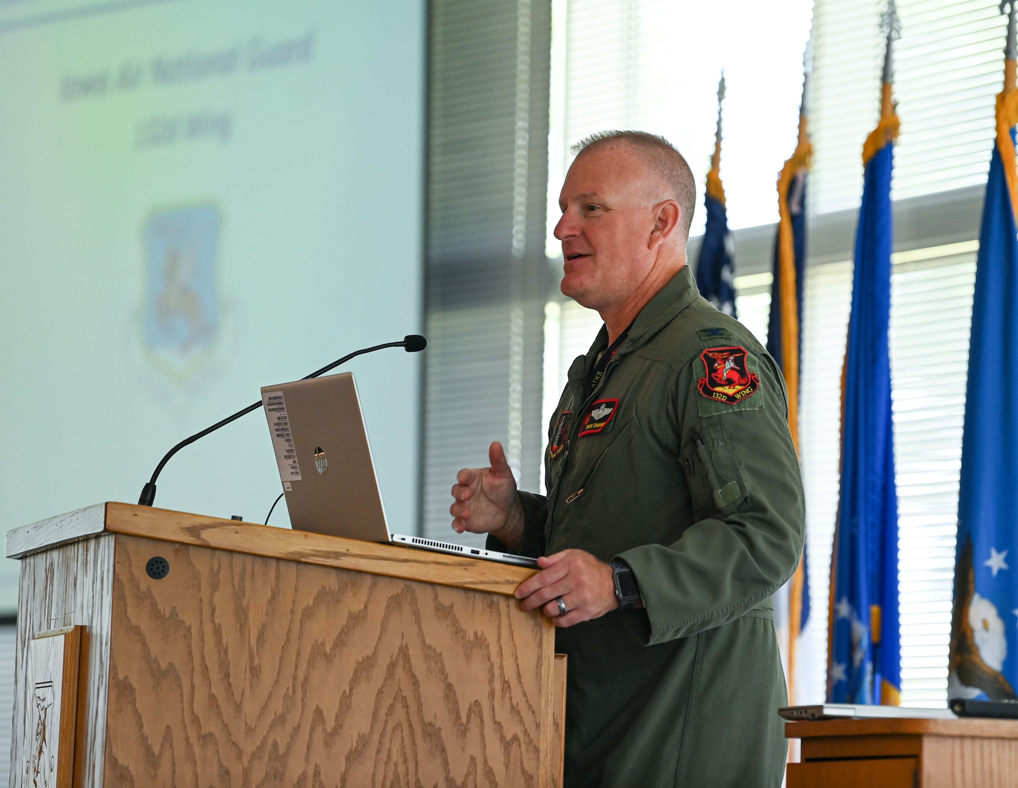 Air Force pilot in green flight suit stands behind podium speaking