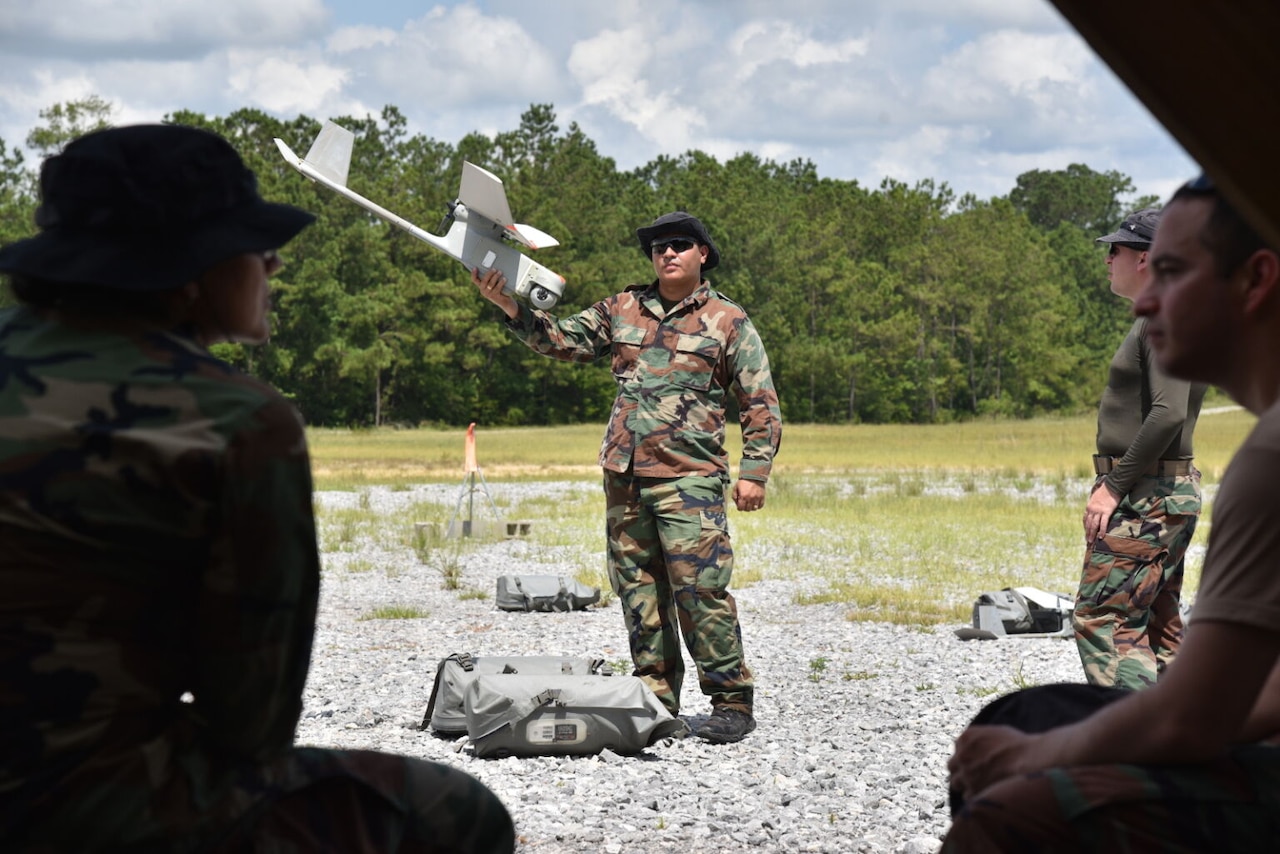 A soldier holds a small drone outside as other soldiers watch.